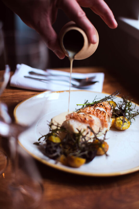 A hand pours sauce over a plated dish featuring fish, garnished with herbs and yellow vegetables, on a wooden table.