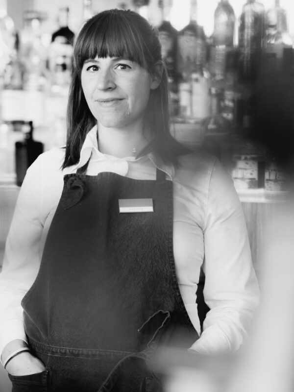 Bartender in a black apron stands confidently in front of a blurred bar background, captured in black and white.