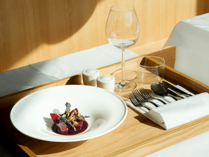 Elegant dish with red beet, garnished, served on a white plate with cutlery and glassware on a wooden tray.