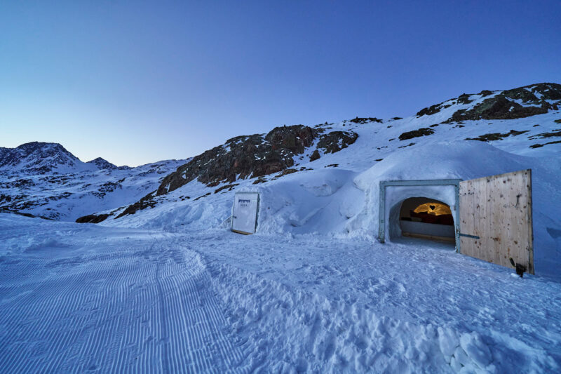 Snow-covered landscape with a wooden hut entrance, surrounded by mountains under a twilight sky.