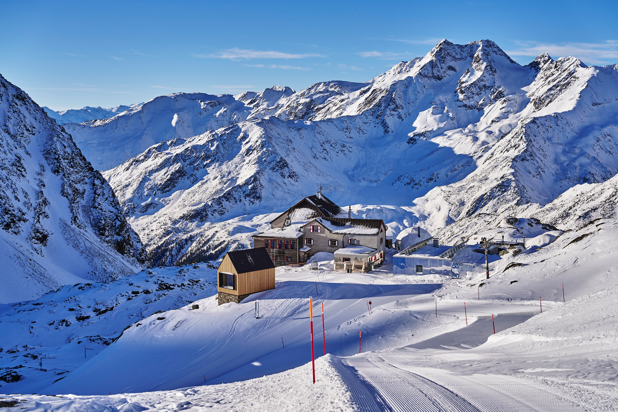Snow-covered mountains surround a chalet in a winter landscape, with ski tracks leading towards the building.