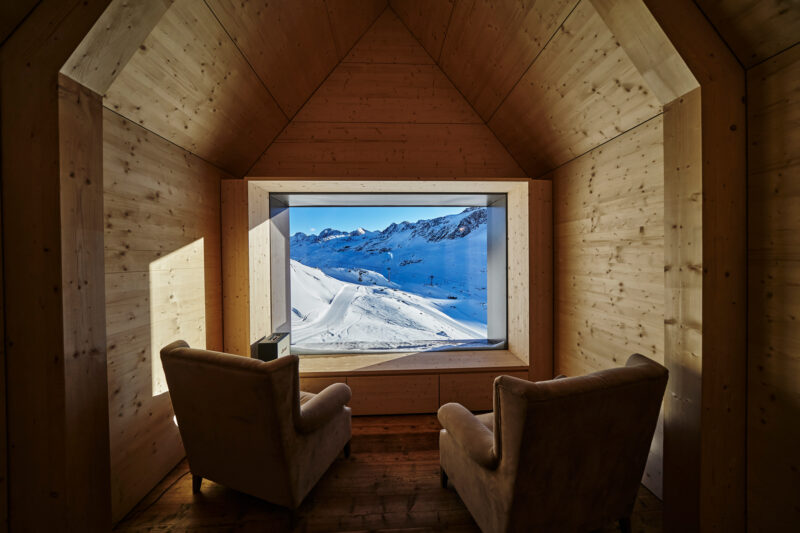 Cozy wooden interior with two armchairs facing a large window showcasing a snowy mountain landscape.