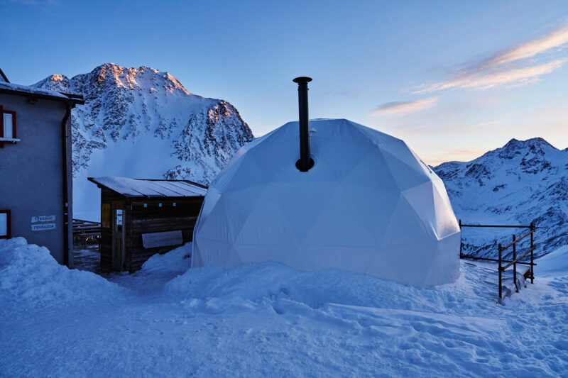 Dome-shaped hut with a chimney, surrounded by snow, against a backdrop of mountains at sunset.