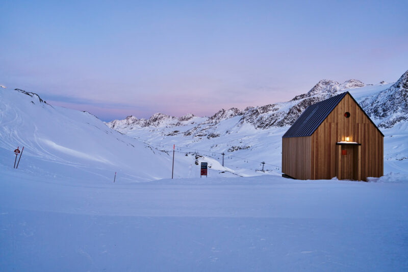 Snow-covered landscape at dusk featuring a wooden hut with a sloped roof, surrounded by mountains.