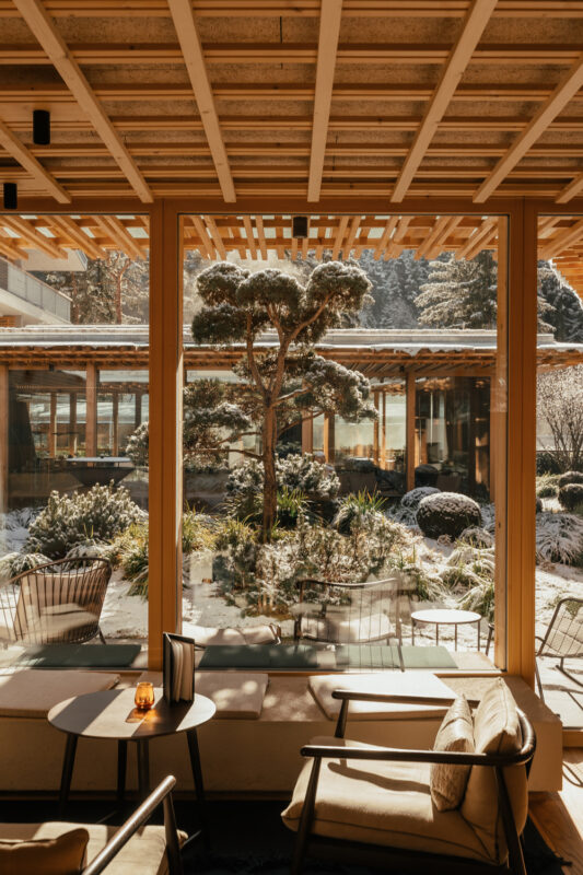 Cozy interior view through large windows, showcasing a snow-covered garden with a bonsai tree and modern seating.