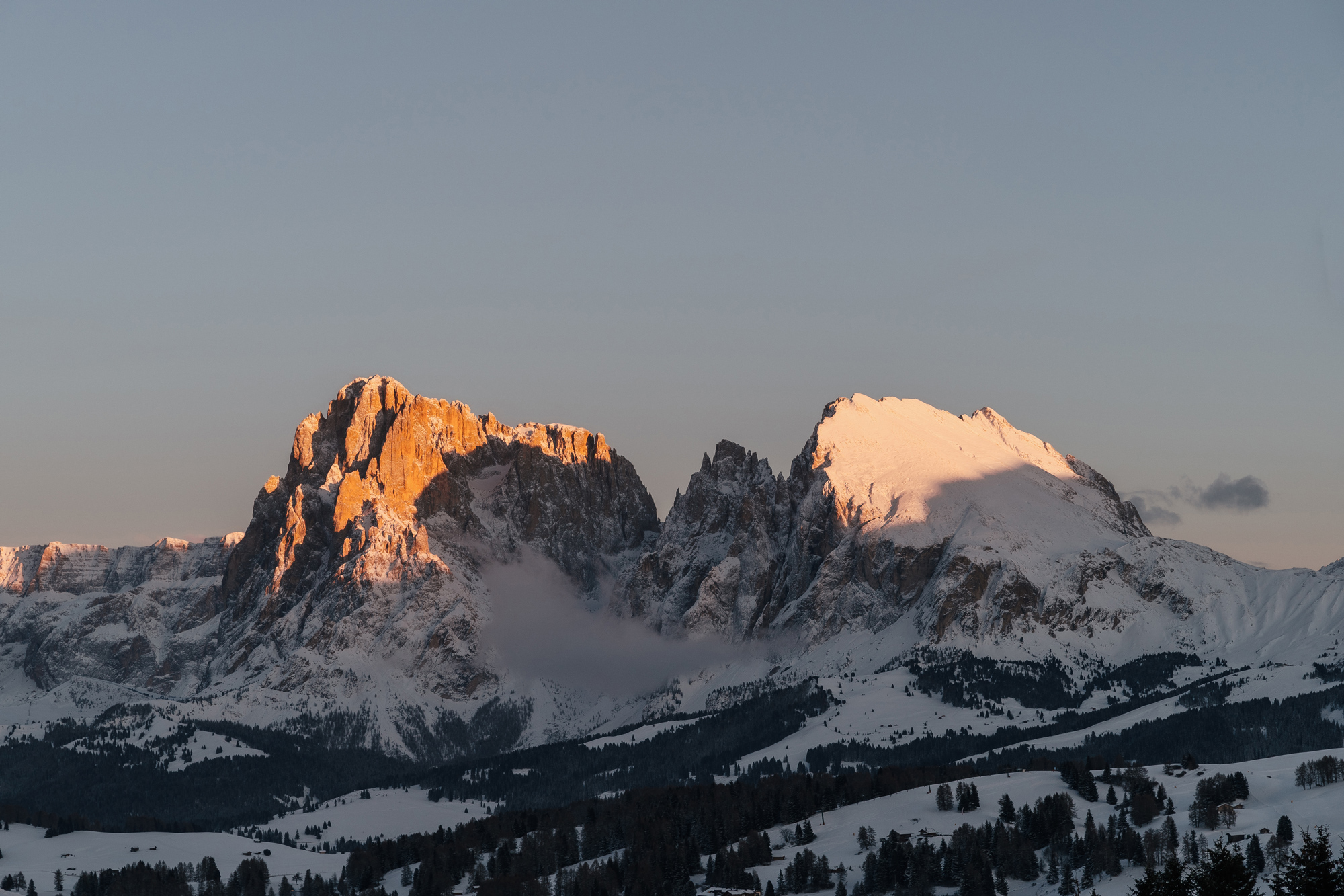 Snow-covered Dolomite mountains at sunset, with warm light illuminating the peaks and a cloudy sky.