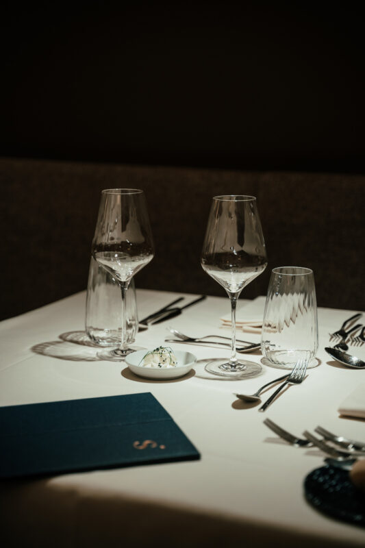 Elegant dining table set with wine glasses, cutlery, and a small dish, featuring a dark, cozy ambiance.