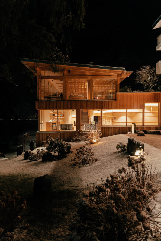 Modern wooden house illuminated at night, surrounded by snow-covered ground and sparse landscaping.