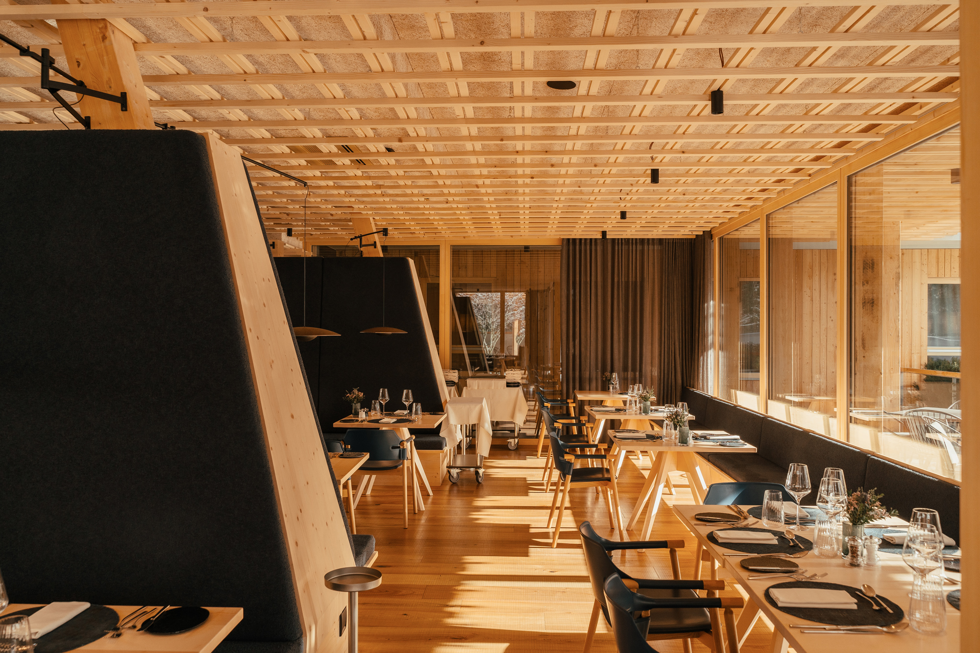 Modern restaurant interior with wooden furnishings, large windows, and neatly set tables, featuring cozy booths.