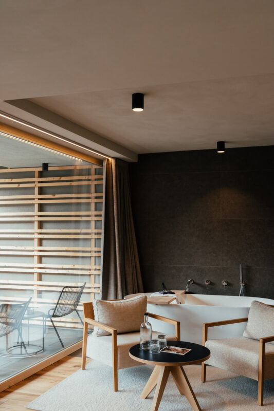 Modern hotel room featuring a bathtub, wooden accents, and a view of a balcony with chairs.
