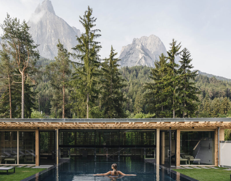 Woman swimming in an outdoor pool surrounded by trees and mountains at Sensoria Dolomites.