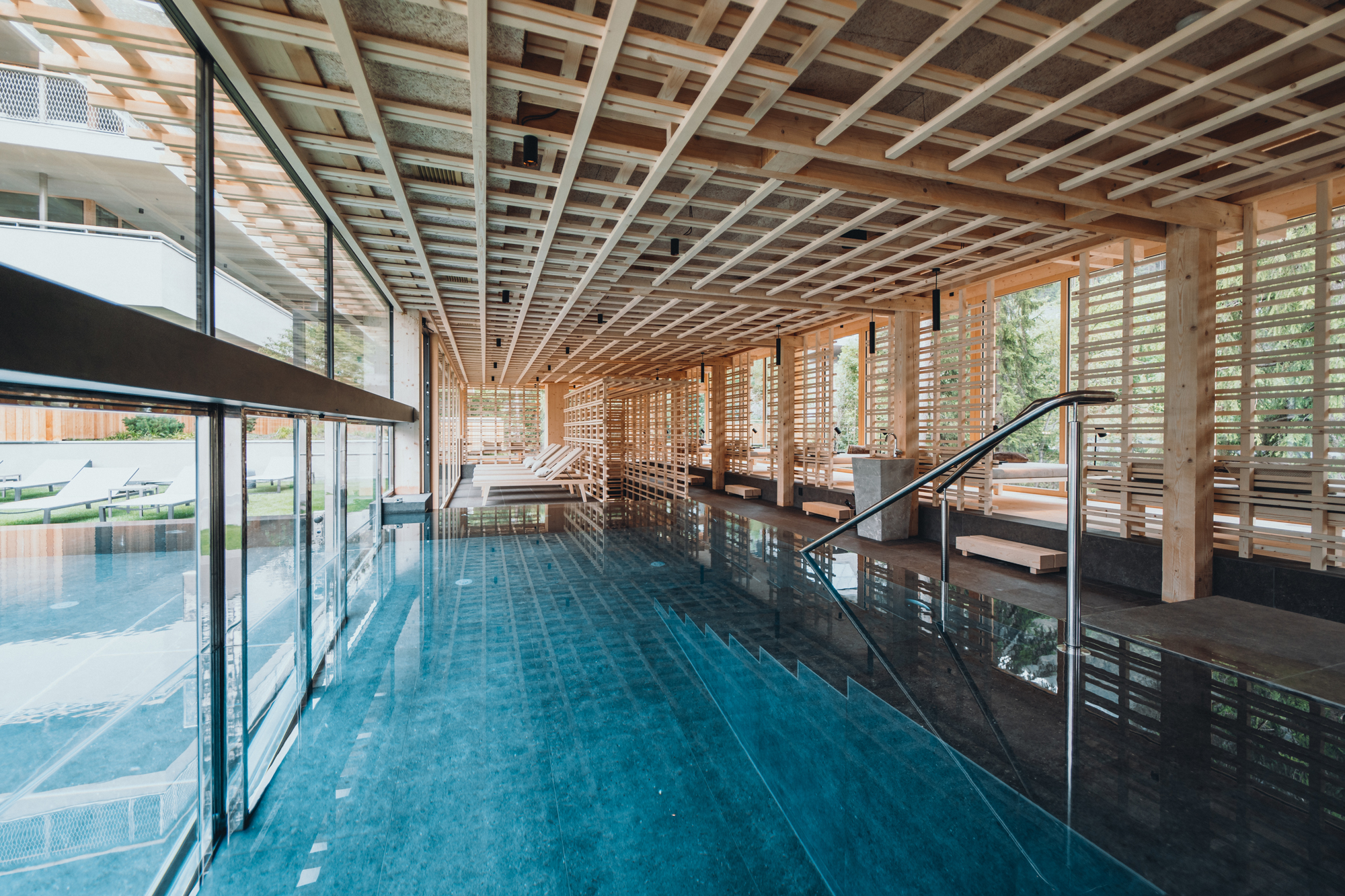 Modern wooden spa interior with a reflective pool and lounge chairs, surrounded by large windows in Sensoria Dolomites.