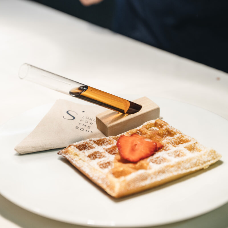 Waffle topped with a strawberry, accompanied by a test tube of sauce on a wooden holder and a napkin.