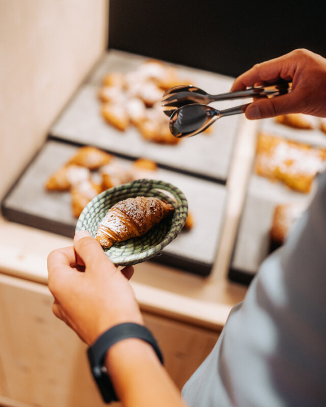 A person holding a green plate with a croissant, tongs in the other hand, near a display of pastries.