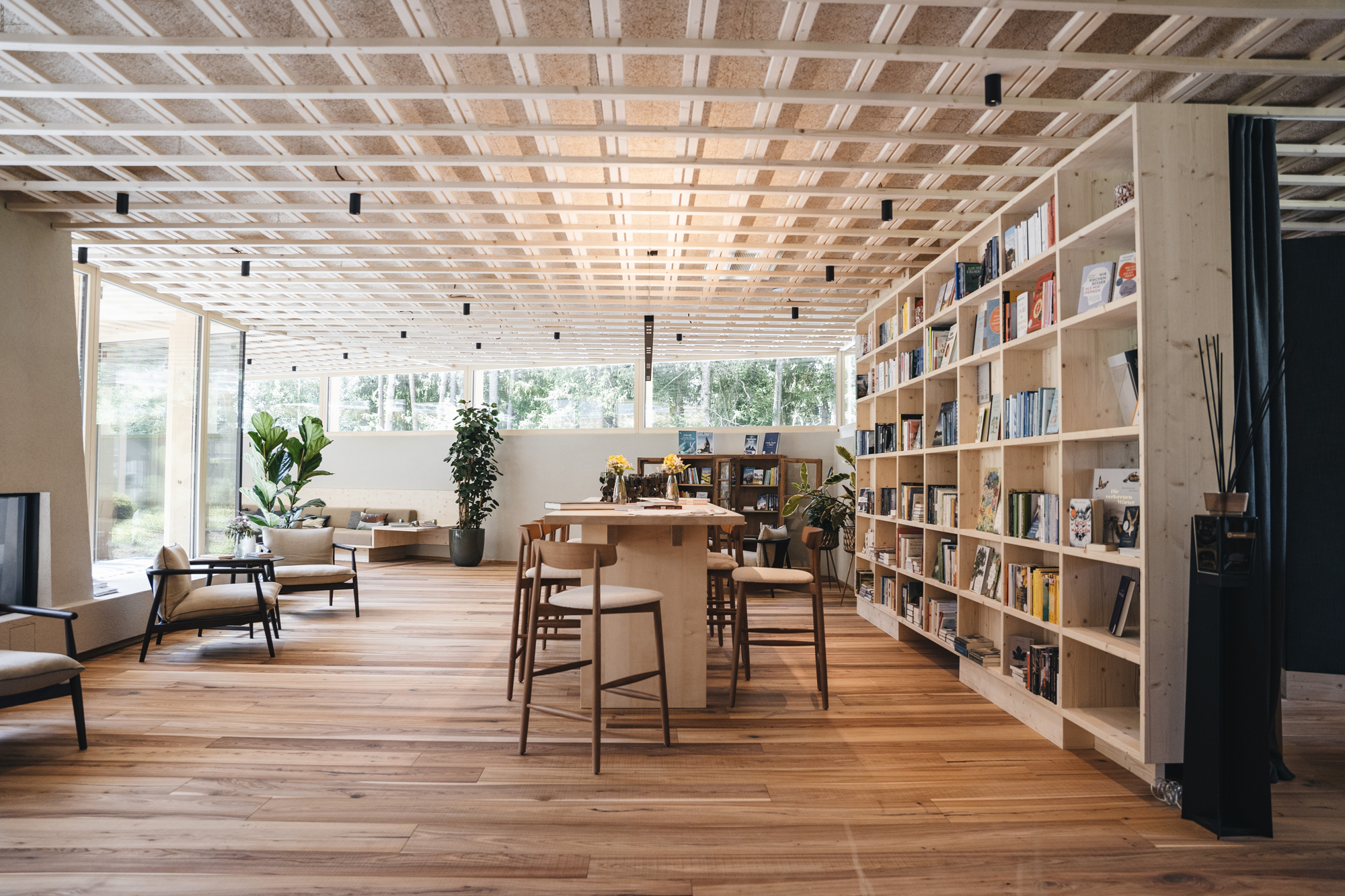 Modern lounge area with wooden flooring, bookshelves, and large windows, featuring plants and seating arrangements.