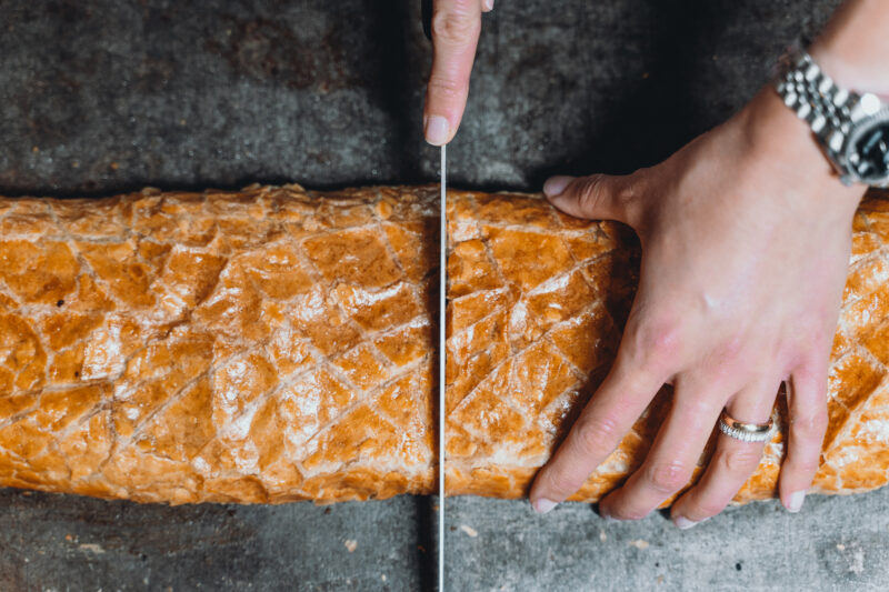 A hand with a ring cuts into a golden-brown pastry on a dark surface, showcasing a textured crust.