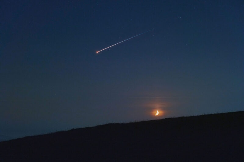 A shooting star streaks across a night sky with a crescent moon visible above a dark horizon.