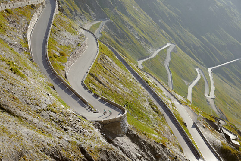 Winding mountain road with cyclists, surrounded by steep green slopes, captured in the Stelvio Pass area.