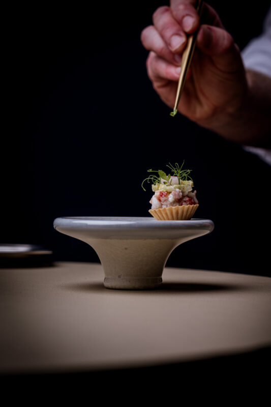 A hand uses chopsticks to garnish a small, elegant dish on a minimalist pedestal against a dark background.