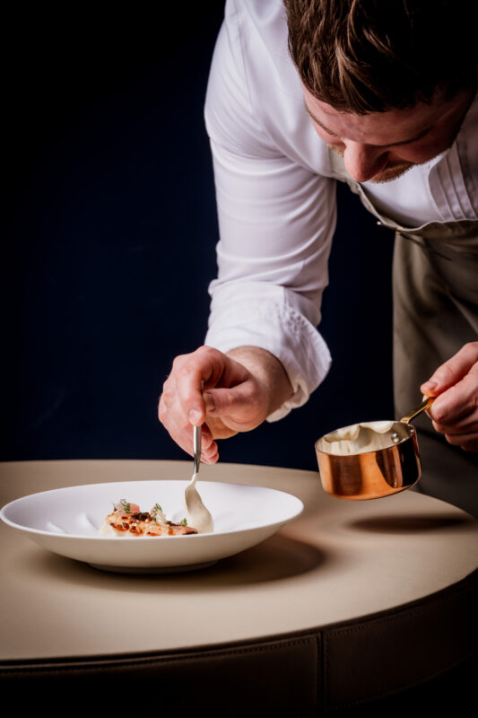 Chef meticulously plating a gourmet dish on a white plate, with a copper sauce ladle in hand, against a dark background.