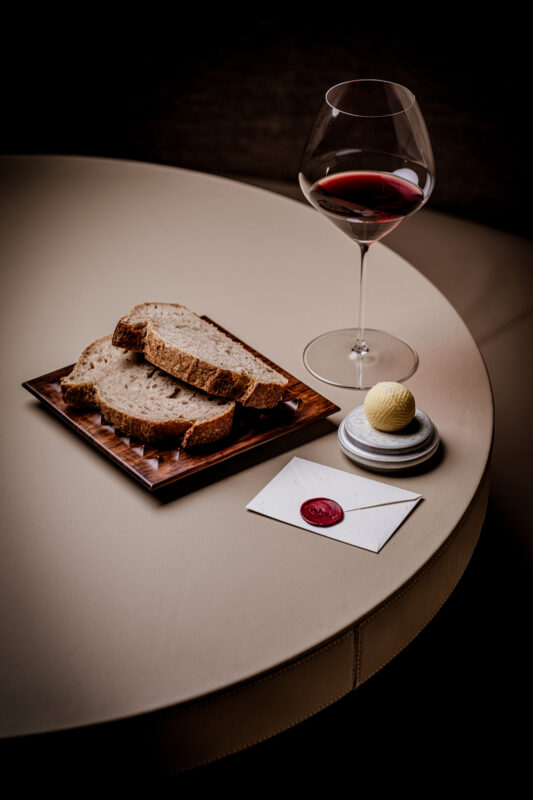 Sliced bread on a wooden plate, a glass of red wine, a small dessert, and a sealed envelope on a round table.