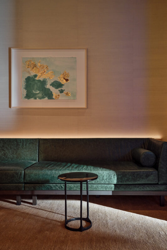 Green velvet sofa with a round black side table, framed artwork above, soft lighting in a modern lobby setting.