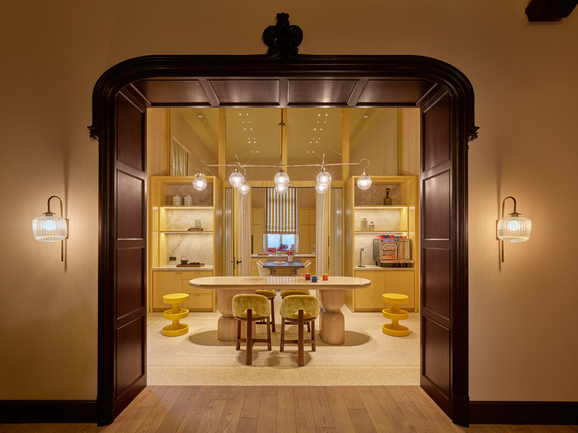 Bright kitchen area with a round table, yellow stools, and pendant lights, framed by an elegant archway.