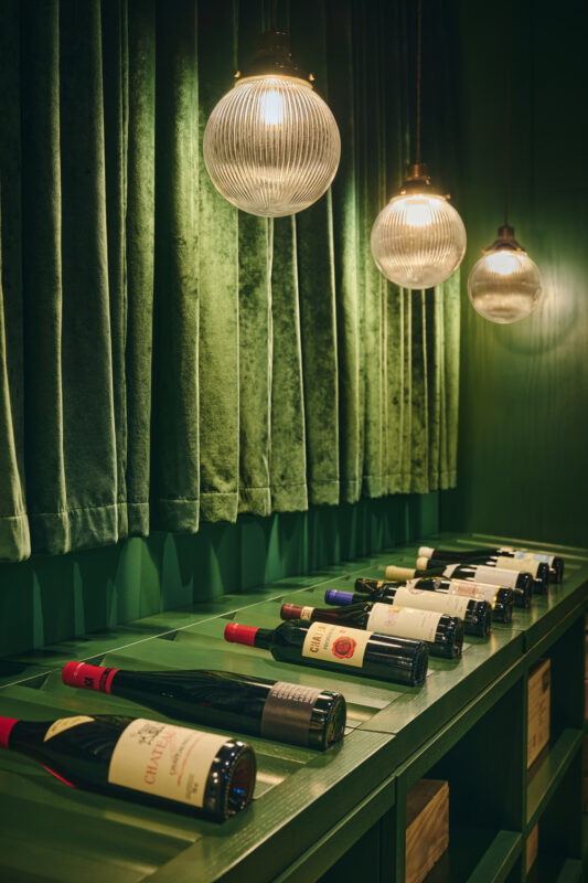 Wine bottles arranged on a green shelf, illuminated by pendant lights, with velvet curtains in the background.