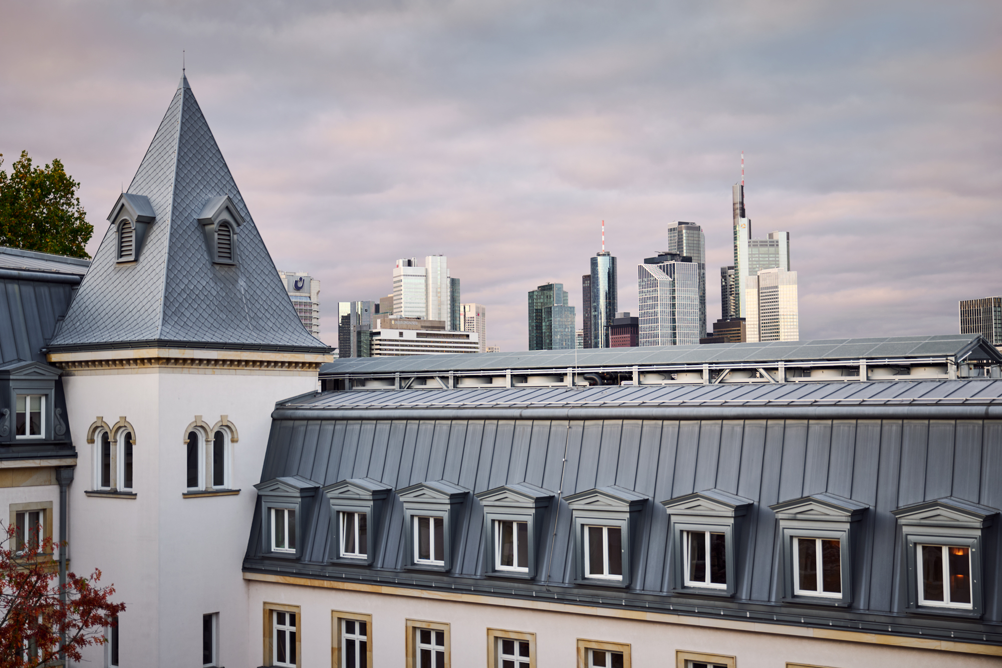 View of a building with a pointed roof and multiple windows, overlooking a city skyline with skyscrapers under a cloudy sky.