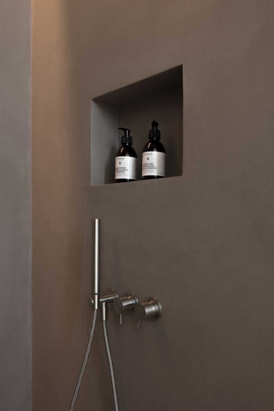 Minimalist bathroom wall with a recessed shelf holding two black bottles, featuring a sleek faucet below.