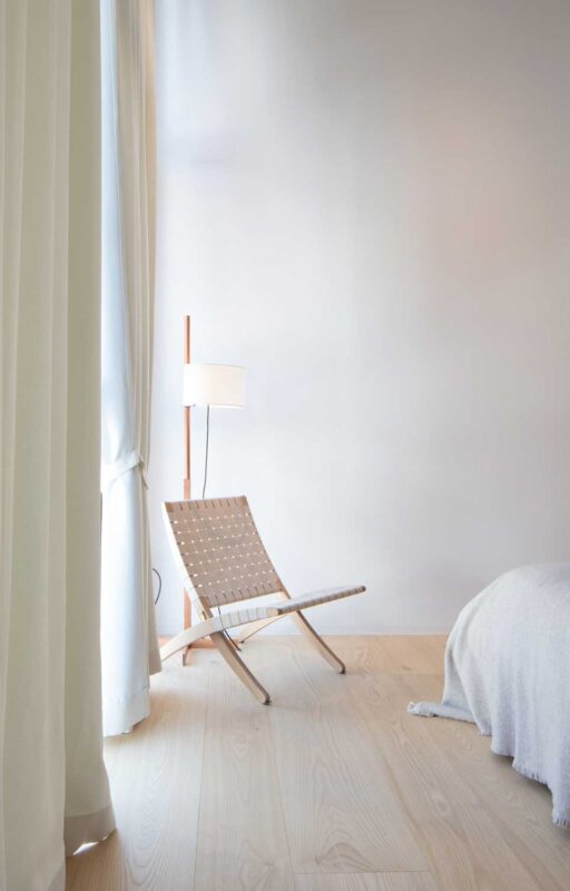 Minimalist bedroom corner featuring a wooden chair, floor lamp, and light curtains, with warm wooden flooring.