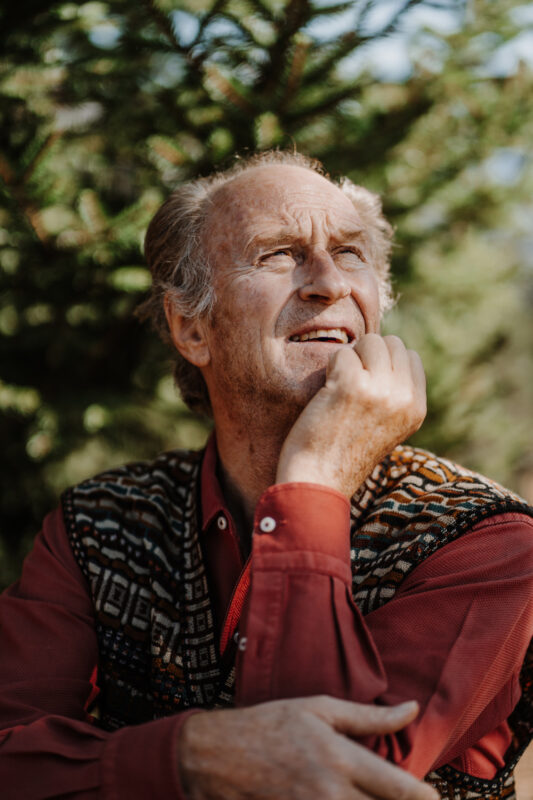 Elderly man in a patterned vest, thoughtfully gazing upwards among greenery.
