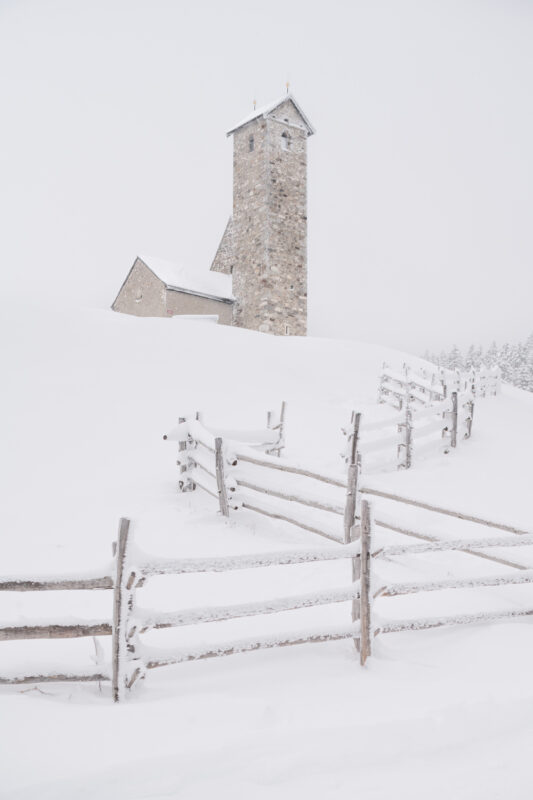 Snow-covered landscape featuring a stone tower and a winding wooden fence in a winter setting.