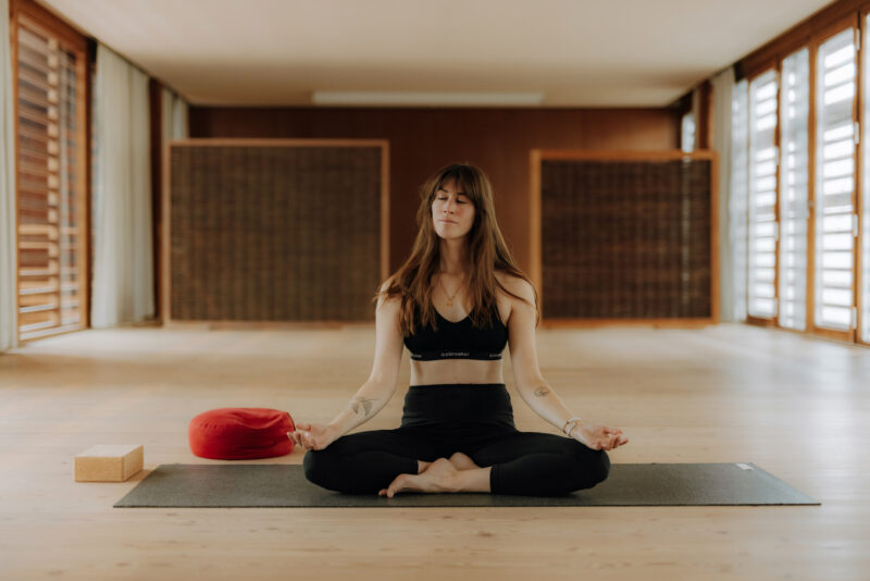 Woman in a black sports bra and leggings meditating on a yoga mat in a serene indoor space with wooden floors.