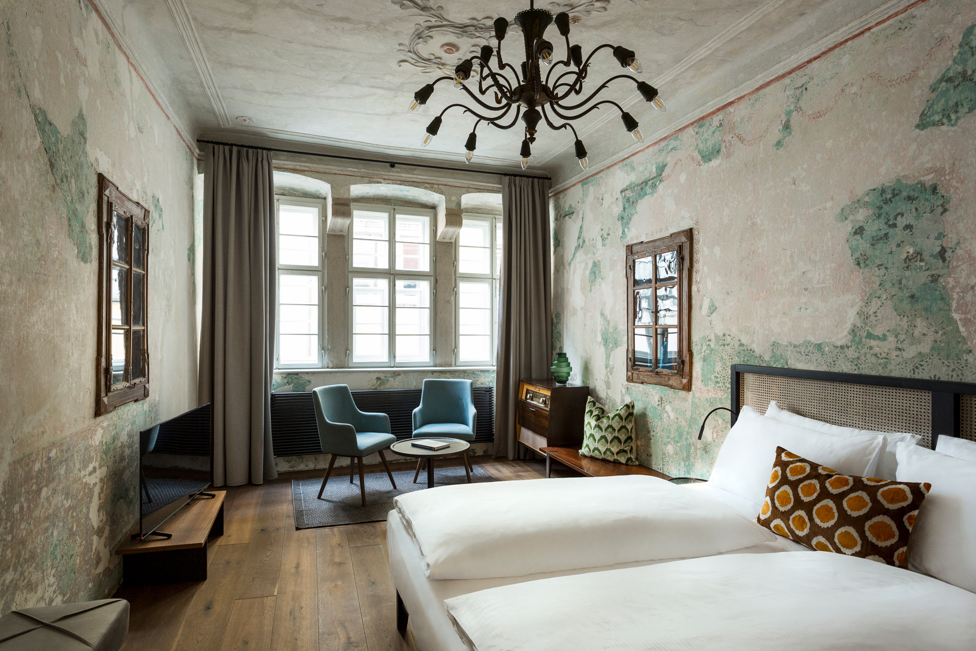 Stylish hotel room with two beds, vintage decor, peeling wall paint, and modern furniture including chairs and a table.