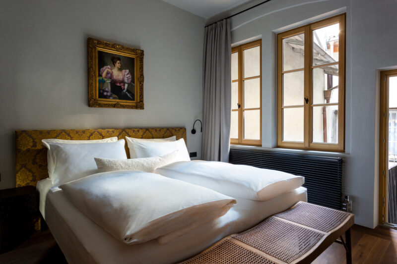 Elegant bedroom featuring a large bed with white linens, a vintage painting above, and large windows letting in natural li...