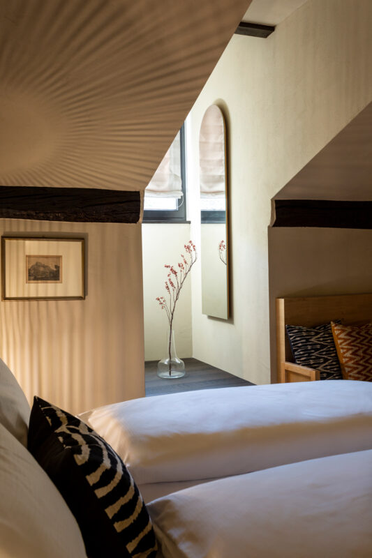 Cozy bedroom with two beds, a decorative vase with flowers, and soft lighting highlighting textured walls.
