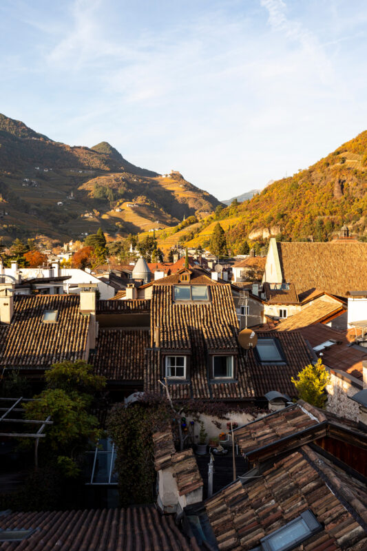 Rooftops of a village with tiled roofs, surrounded by mountains and autumn foliage under a clear sky.
