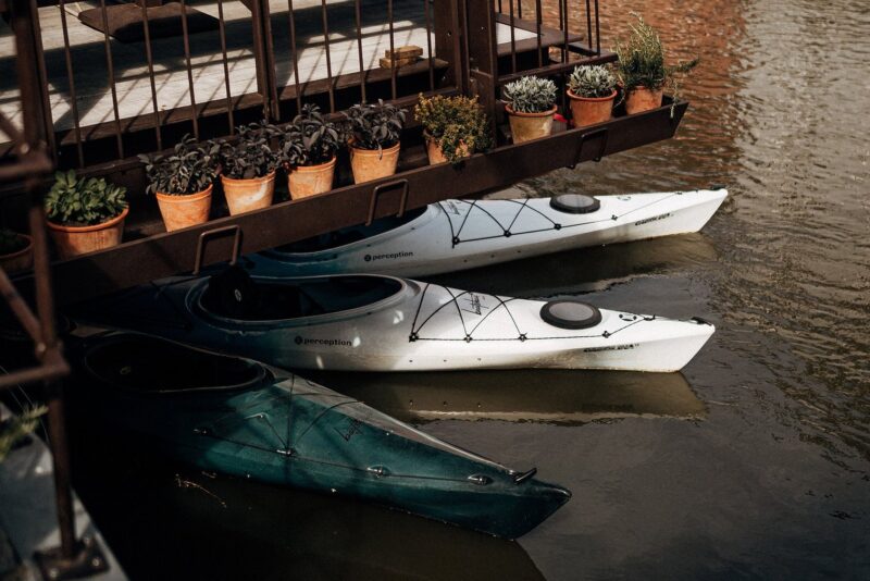 Three kayaks, one green and two white, are moored beneath a deck with potted plants along the edge.