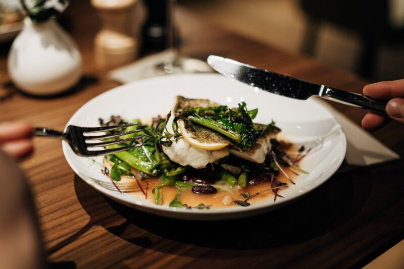 Plate of fish topped with greens and lemon, garnished with microgreens, on a wooden table.