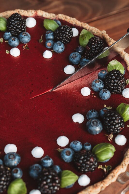 Red berry tart topped with blueberries, blackberries, and mint, with a knife cutting into the dessert.
