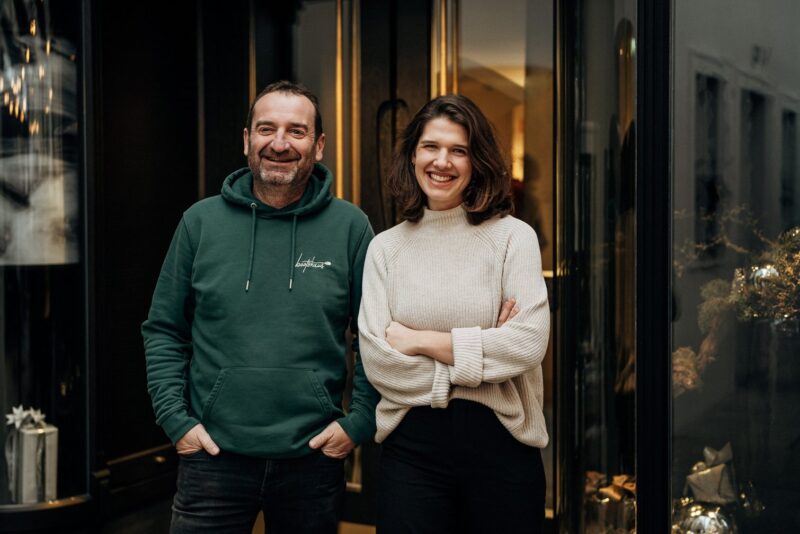 Man and woman smiling, standing together in front of a glass entrance, wearing casual attire.