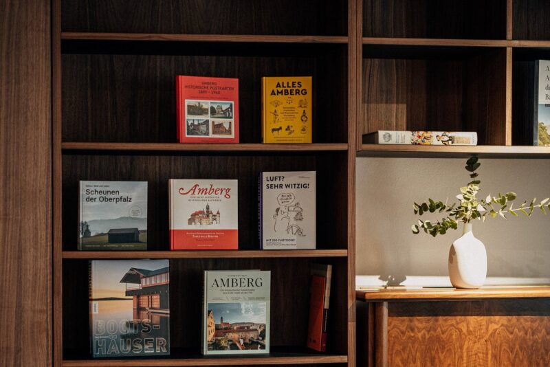 Books displayed on wooden shelves, featuring titles about Amberg and Oberpfalz, with a decorative vase on the right.