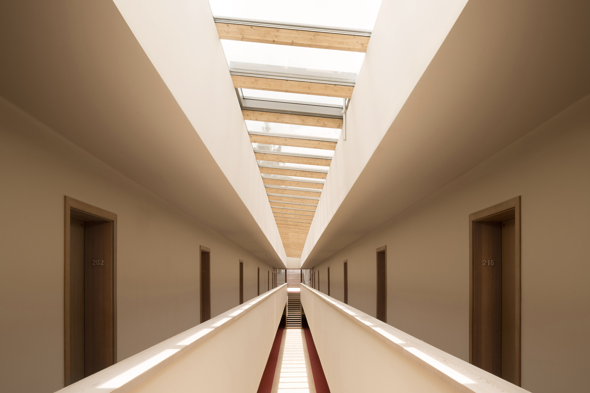 Long corridor with wooden doors numbered 202 and 216, skylights above, and a red runner leading to the end.
