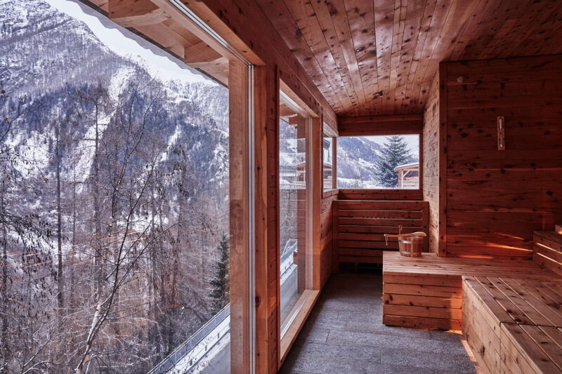 Wooden sauna interior with large windows showcasing a snowy mountain landscape outside.