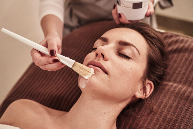 A woman receiving a facial treatment, with a hand applying a cream using a brush.