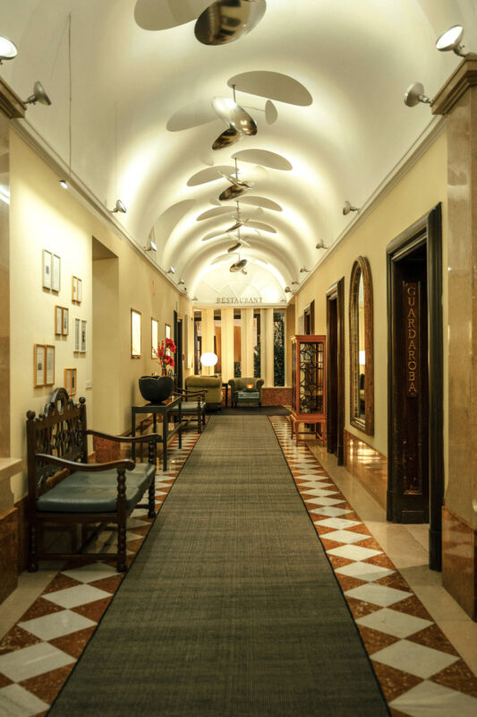 Elegant hotel corridor with a patterned floor, decorative lighting, and seating areas, leading to a restaurant.
