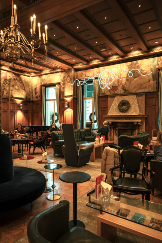 Elegant hotel lounge with ornate decor, plush seating, and a grand chandelier, featuring guests relaxing and a piano.