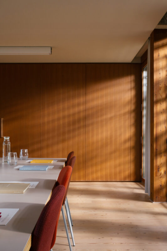 Bright meeting room with a wooden wall, a long table set with glassware, and sunlight casting shadows on the floor.
