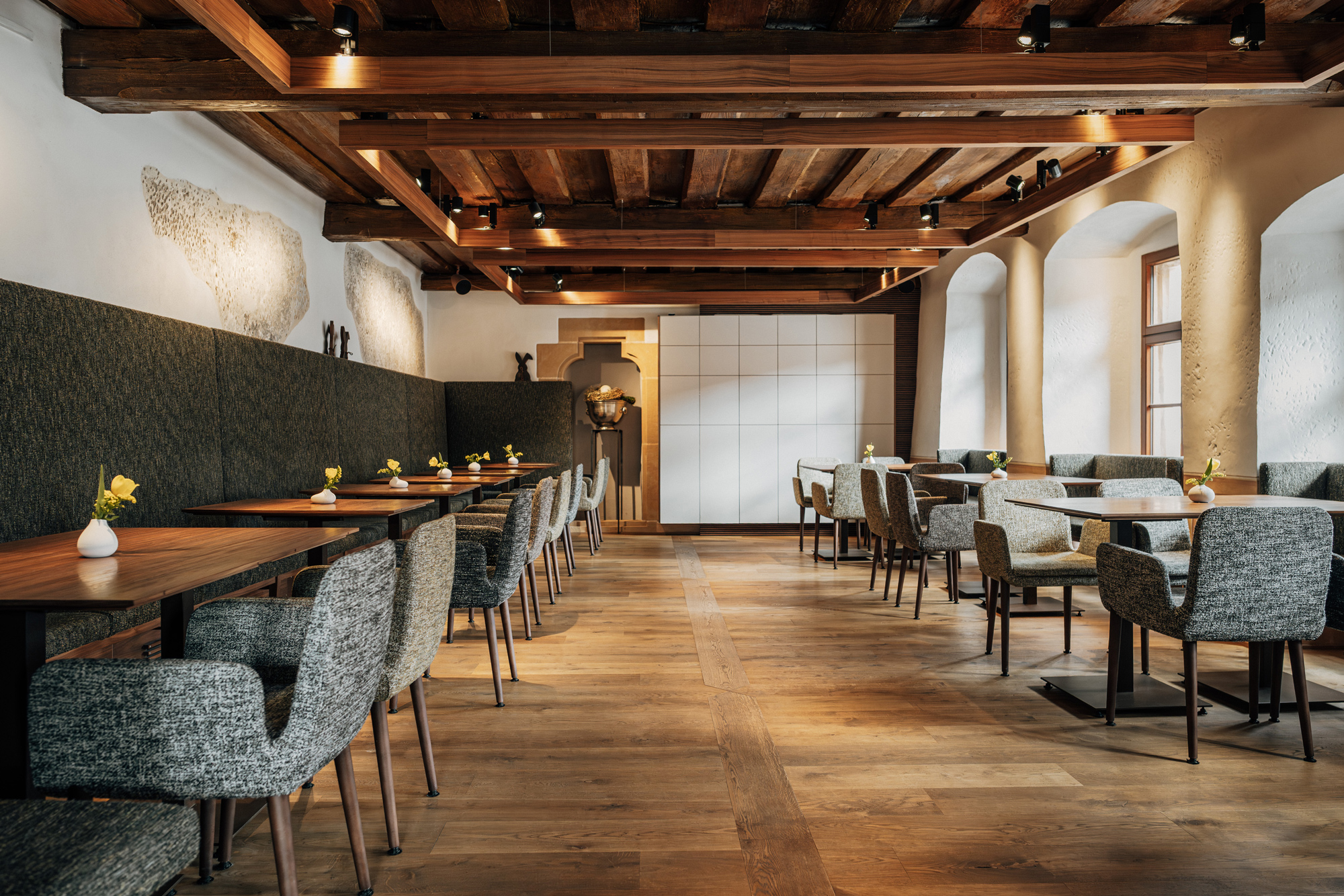 Modern restaurant interior with wooden beams, tables set with flowers, and cozy seating arrangements.